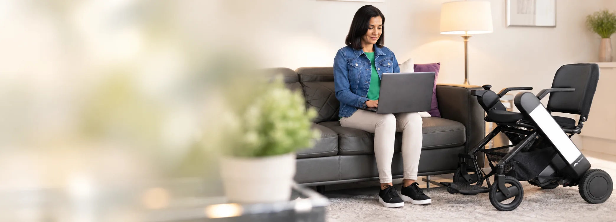 A woman working on a laptop at home with her WHILL power wheelchair beside the sofa